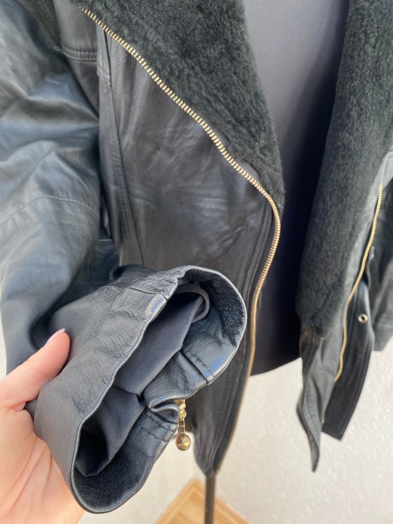 K&ouml;nnte beinhalten: Schwarze Lederjacke mit Rei&szlig;verschluss und Pelzkragen. Die Jacke hat eine Rei&szlig;verschlusstasche auf der linken Seite und einen goldenen Rei&szlig;verschluss.