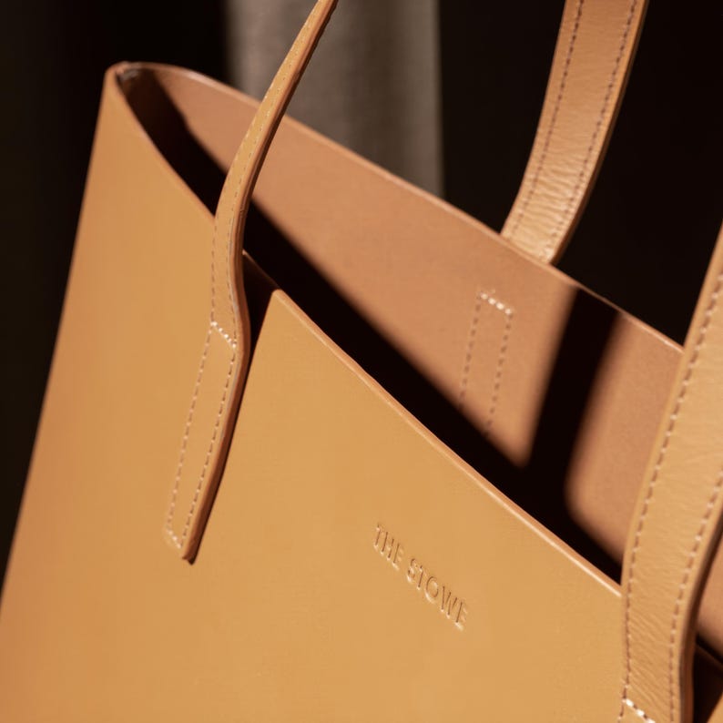 Puede incluir: Un bolso tote de cuero marr&oacute;n con el texto "THE  STOWE" grabado en la parte delantera.