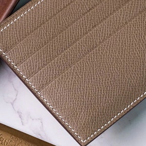 May include: A close-up of a tan leather cardholder with white stitching. The cardholder has multiple card slots and a textured surface. The edges are neatly finished, and the overall design is minimalist and elegant.