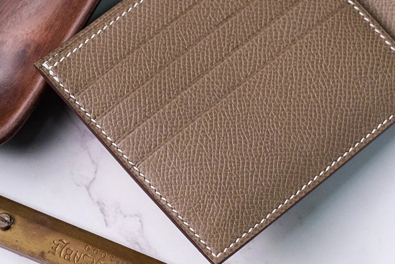 May include: A close-up of a tan leather cardholder with white stitching. The cardholder has multiple card slots and a textured surface. The edges are neatly finished, and the overall design is minimalist and elegant.