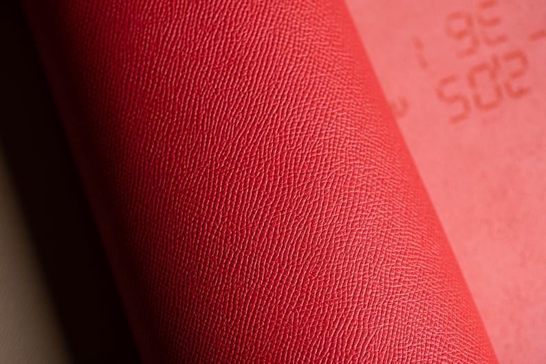 May include: Close-up of a rolled red leather material with a textured surface. The material is a vibrant shade of red, with a subtle pattern visible on the surface. The roll is partially visible, with the edge of the material showing.