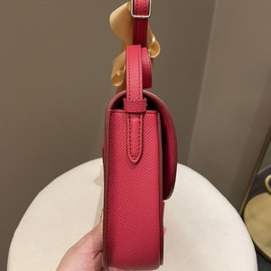 May include: A side view of a red leather crossbody bag with a flap closure and adjustable strap. The bag has a silver buckle and a textured surface. A light-colored ribbon is attached to the strap.
