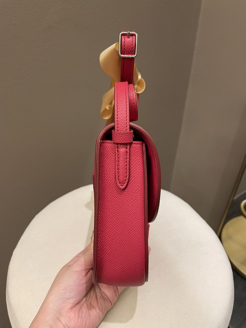 May include: A side view of a red leather crossbody bag with a flap closure and adjustable strap. The bag has a silver buckle and a textured surface. A light-colored ribbon is attached to the strap.