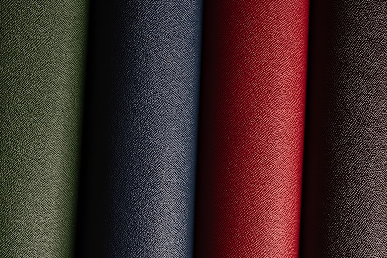 May include: Close-up of four rolls of textured material in shades of green, navy blue, red, and brown. The material has a subtle, repeating pattern, suggesting a leather or similar fabric. The rolls are tightly wound, showcasing the material's texture.