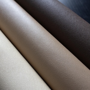 Puede incluir: Primer plano de tres rollos de cuero sintético texturizado en diferentes tonos. Los colores varían desde un crema claro hasta un beige medio y un marrón oscuro. El material tiene un grano sutil y granulado, lo que sugiere un acabado de alta calidad.