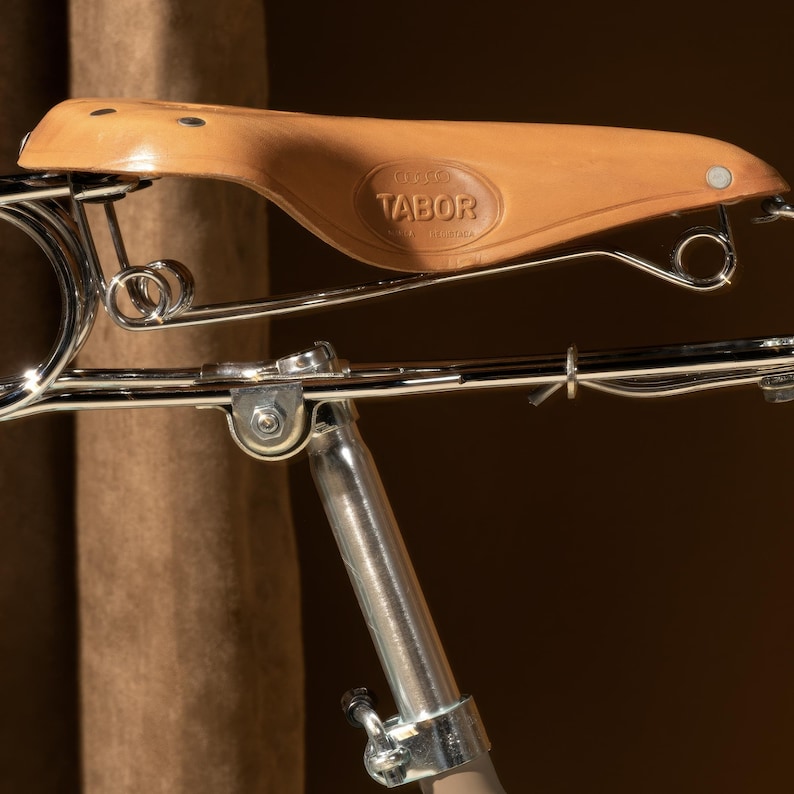 Puede incluir: Primer plano de un sill&iacute;n de bicicleta de cuero marr&oacute;n con un marco cromado y un logotipo de Tabor met&aacute;lico. El sill&iacute;n est&aacute; sujeto a un poste de bicicleta cromado.