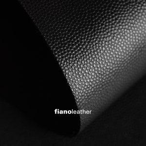 Pode incluir: Close-up de couro preto texturizado com um brilho sutil. O couro está dobrado, criando uma forma curva. O texto "fianoleather" é visível no canto inferior direito.