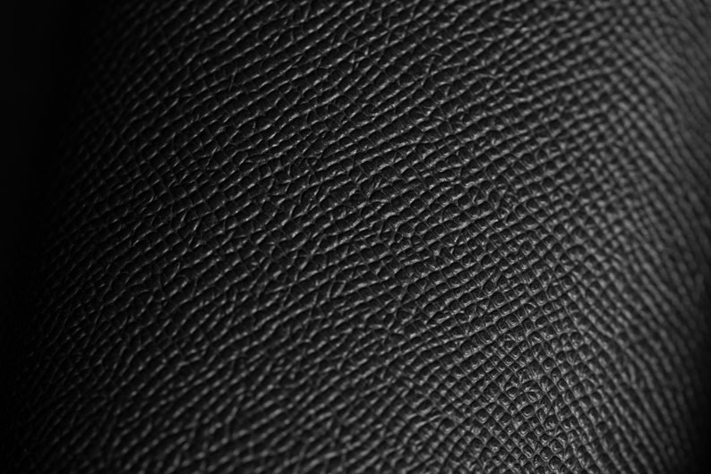 May include: Close-up of black leather with a textured surface. The leather has a pattern of small, irregular shapes and lines, creating a detailed and tactile appearance. The lighting highlights the texture, giving it depth and dimension.