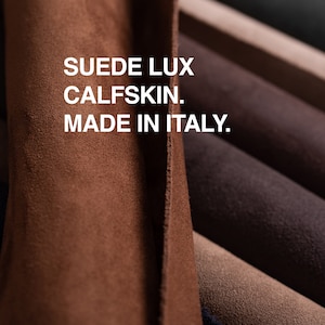 May include: Close-up of a stack of brown, beige, and gray suede leather swatches. The text "SUEDE LUX CALFSKIN. MADE IN ITALY." is printed on the top swatch.