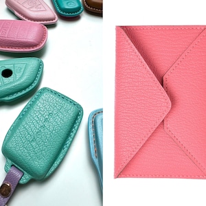 May include: Assortment of colorful leather key covers in pink, teal, and blue. The covers are made of textured leather and have a stitched edge. A pink leather envelope style pouch is also shown.