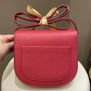 May include: A red crossbody bag with a rounded flap and a matching adjustable strap. The bag features a small exterior pocket and is adorned with a gold ribbon. The bag's dimensions are not visible in the image.