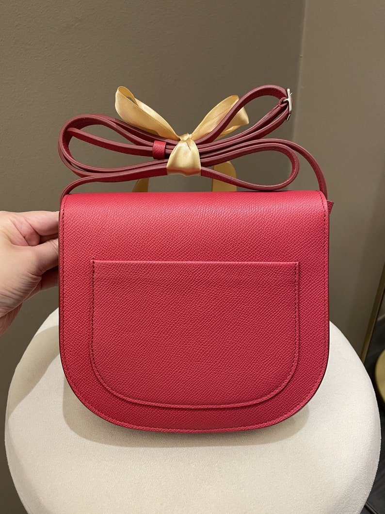 May include: A red crossbody bag with a rounded flap and a matching adjustable strap. The bag features a small exterior pocket and is adorned with a gold ribbon. The bag's dimensions are not visible in the image.