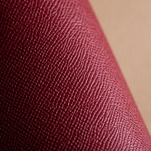 May include: Close-up of a rolled section of burgundy leather. The leather has a textured, cross-hatched pattern. The background is a light tan color, providing contrast to the rich, dark red material. The image highlights the texture and color of the leather.