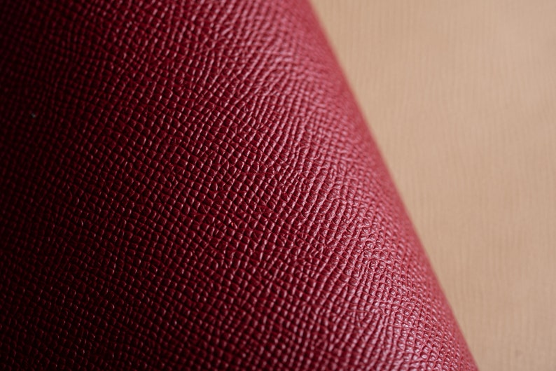May include: Close-up of a rolled section of burgundy leather. The leather has a textured, cross-hatched pattern. The background is a light tan color, providing contrast to the rich, dark red material. The image highlights the texture and color of the leather.