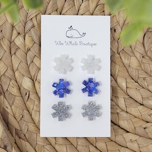May include: A white card displays six snowflake-shaped earrings in three different colors: white, blue, and silver. The earrings are arranged in pairs. The card has the text "Wee Whate Boutique" and a whale logo.
