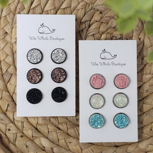 May include: Two sets of stud earrings on white cards. One set features silver, rose gold, and black glitter studs. The other set includes pink, white, and blue glitter studs. Each card has the text "Wee Whale Boutique" and a whale logo.