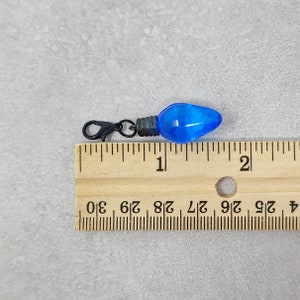 May include: A blue light bulb charm with a black metal clasp. The charm is about 1 inch long and is shown next to a ruler.