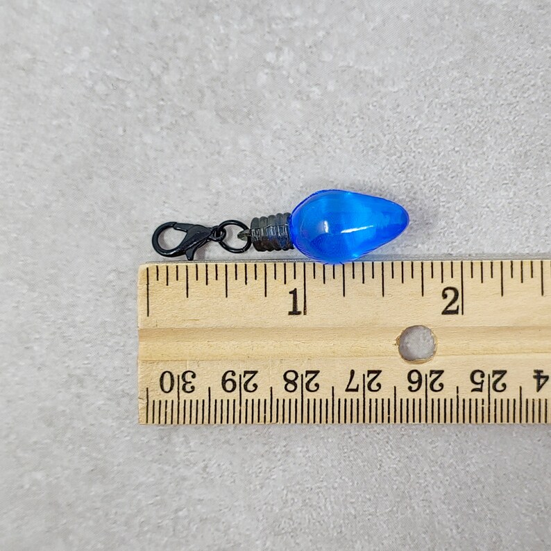 May include: A blue light bulb charm with a black metal clasp. The charm is about 1 inch long and is shown next to a ruler.