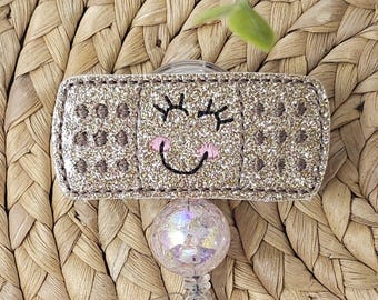 Op de afbeelding: Een glitter badge reel met een lachend gezichtsontwerp. De rechthoekige badge heeft een gouden glitter afwerking met geborduurde ogen en een roze mond. De reel heeft een helder, iriserend, rond ontwerp.