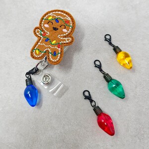 May include: A gingerbread man with a smiling face and red, green, and yellow lights is attached to a retractable badge holder. Three light bulb charms are included in blue, green, and red.
