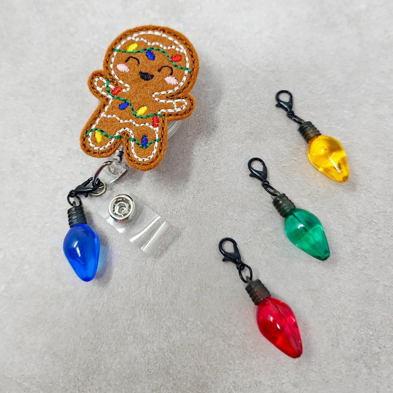 May include: A gingerbread man with a smiling face and red, green, and yellow lights is attached to a retractable badge holder. Three light bulb charms are included in blue, green, and red.