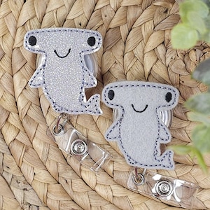 May include: Two hammerhead shark-shaped badge reels. One is silver glitter, the other is light gray felt. Both have black eyes and a stitched smile. They are attached to clear plastic reels with metal clips.