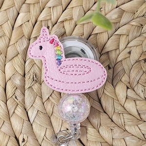 Op de afbeelding: Een badgehouder met een lichtroze eenhoorn-zwemband van kunstleer met een regenboogkleurige manen. De eenhoorn is bevestigd aan een transparante plastic haspelbehuizing en een bolvormige kraal met een glinsterende, iriserende afwerking.