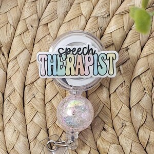 May include: A badge reel with the words "Speech Therapist" in pastel colours. The reel has a clear plastic casing and a glittery, iridescent ball at the bottom. The background is a woven, textured surface.