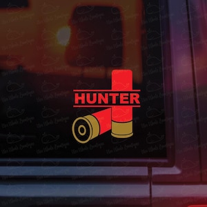 May include: A red and gold graphic featuring two shotgun shells and the word "HUNTER" in red capital letters. The design is likely a decal or sticker, with a simple, bold aesthetic.