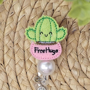 May include: A light green felt cactus with a smiling face and embroidered details sits in a pink pot that reads "Free Hugs." The badge reel has a white pearl bead and a silver clip.