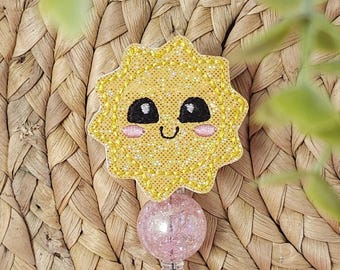May include: A yellow embroidered badge reel featuring a smiling sun design with black eyes and a pink blush. The badge reel has a glittery pink ball and a silver clip. The background is a woven texture.