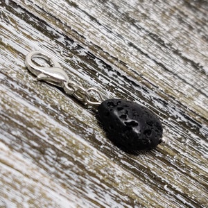 Puede incluir: Un charm plateado con una cuenta de roca de lava negra. El charm tiene un cierre de langosta.