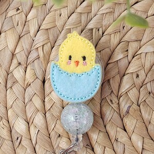 May include: A yellow felt chick hatching from a light blue egg, attached to a clear, glittery badge reel. The chick has embroidered details. The badge reel has a silver clip.