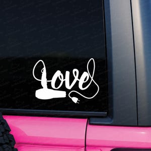 May include: A white vinyl decal applied to a dark window, featuring the word love in a cursive script font. The letter l is shaped like a hair dryer, and the letter e extends into a long, looping cord that ends with a two-pronged electrical plug.