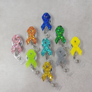 Op de afbeelding: Assortiment van tien kleurrijke glitterlint-badge-rollen. Elke rol heeft een ander gekleurd lint met een glitterafwerking. De linten zijn bevestigd aan een zilveren metalen badge-rol.