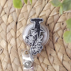 May include: A clear, round badge reel featuring a black glitter hammerhead shark design with white floral accents. The badge reel has a clear plastic clip and a retractable cord.