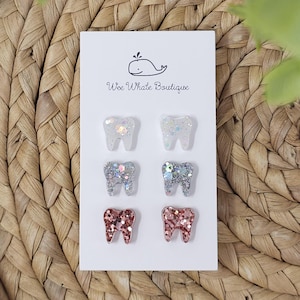 May include: Six tooth-shaped earrings in three different glittery colors: clear, silver, and rose gold. The earrings are displayed on a white card with the text "Wee Whale Boutique" and a whale illustration. The background is a woven wicker texture.