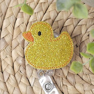 Könnte beinhalten: Ein gelbes, glitzerndes Enten-Abzeichen mit einem orangefarbenen Schnabel und einem schwarzen Auge. Das Abzeichen hat ein durchsichtiges, ausziehbares Kunststoffkabel und einen Metallclip. Die Ente ist mit orangefarbenem Faden genäht.