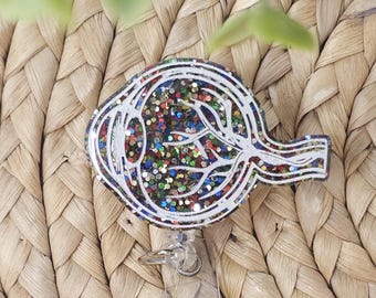 Pode incluir: Um porta-crachá com um diagrama detalhado do olho, contornos brancos e preenchimento de glitter colorido. O porta-crachá tem um cordão retrátil de plástico transparente e um clipe de metal. O fundo é uma superfície tecida e texturizada.
