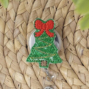 May include: A Christmas tree-shaped badge reel with a red bow at the top. The tree is green with gold string lights and a brown trunk. The badge reel has a clear, round base and a retractable cord with a metal clip.