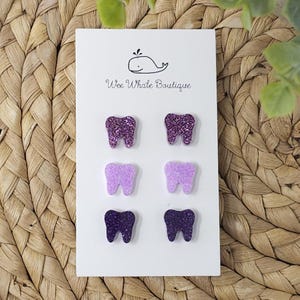 May include: Six tooth-shaped earrings in shades of purple, arranged on a white card. The earrings are glittery and vary in color from dark purple to light lavender. The card has a whale logo and the text "Wee Whale Boutique".