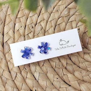 May include: A pair of blue snowflake-shaped earrings with glitter accents, displayed on a white card with the "Wee Whale Boutique" logo. The earrings are set against a woven wicker background.