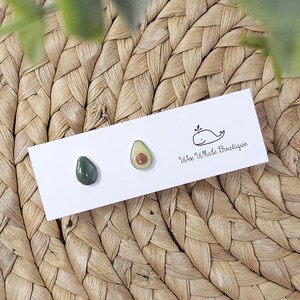 May include: A pair of avocado-themed stud earrings and a single dark green stud earring on a white card. The avocado earrings are green with a brown center. The card has a whale logo and the text "Wee Whale Boutique".