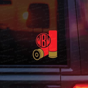 Puede incluir: Una pegatina de cartucho de escopeta roja y dorada con un monograma. La pegatina presenta dos cartuchos rojos y un casquillo dorado. También se incluye un monograma circular con las letras "WBD".