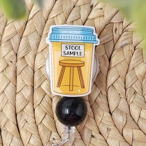 May include: A novelty badge reel featuring a cartoon illustration of a stool sample container. The container is yellow with a blue lid and the words "STOOL SAMPLE" printed on it. A small stool is drawn on the container. The reel has a black, round base.