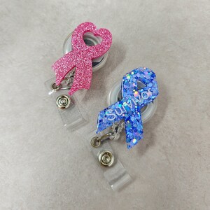 Op de afbeelding: Twee glittereende lint-uittrekbare badgehouders. E&eacute;n is roze met een hartvorm en de andere is blauw met het woord "Survivor" erop.