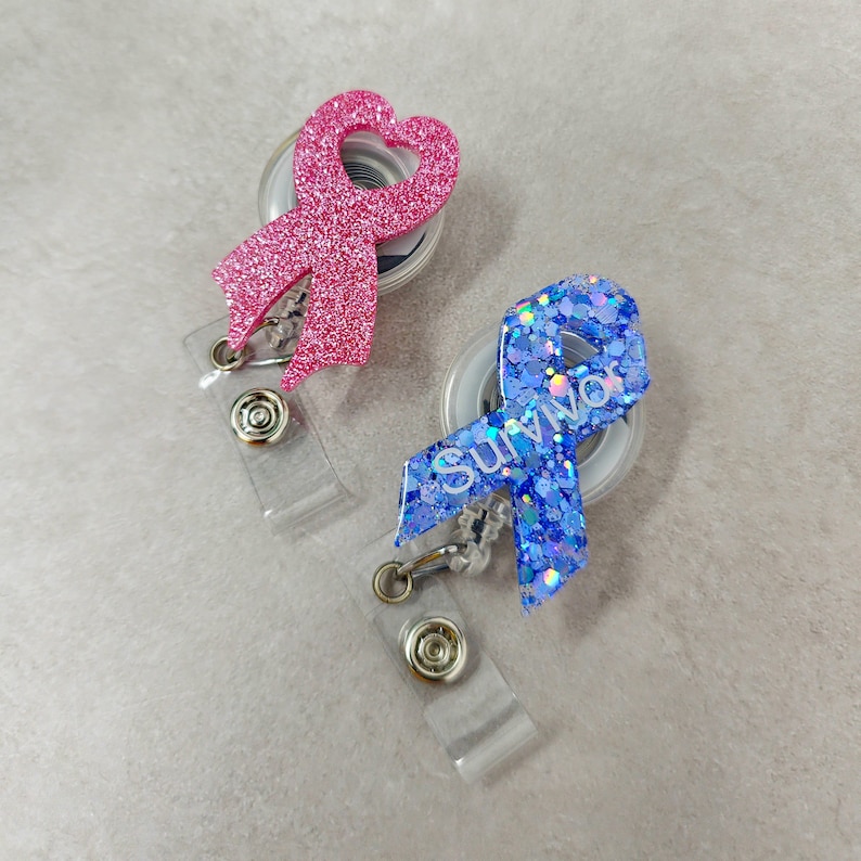 Op de afbeelding: Twee glittereende lint-uittrekbare badgehouders. E&eacute;n is roze met een hartvorm en de andere is blauw met het woord "Survivor" erop.