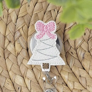 May include: A white Christmas tree-shaped badge reel with a pink bow at the top. The tree has silver stitching and a brown base. The reel is attached to a silver clip and is set against a woven background.