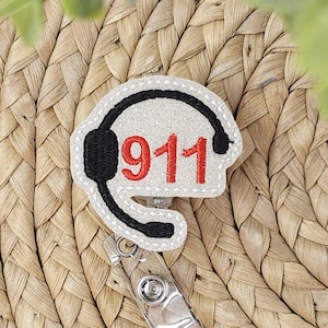 Puede incluir: Un portatarjetas blanco y brillante con un diseño de auriculares negros y el número rojo "911". El portatarjetas tiene un clip plateado y está sobre un fondo tejido.