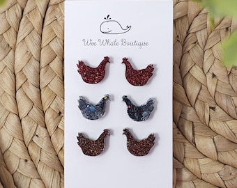 May include: A set of six glittery chicken-shaped earrings in shades of red, black, and brown, displayed on a white card. The card features a whale logo and the text "Wee Whale Boutique". The earrings are arranged in two rows of three.